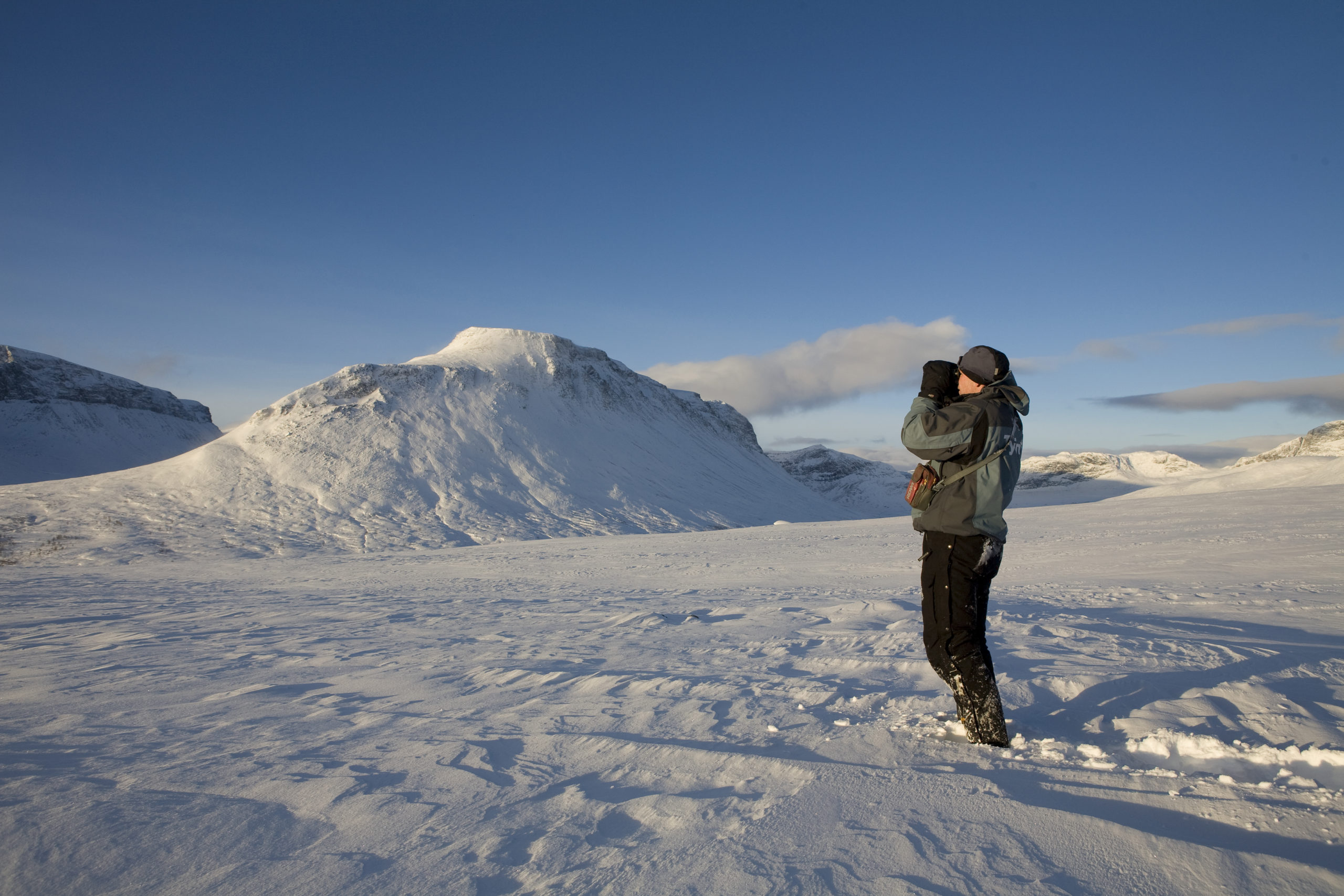 Reingjetar som kikar etter reinflokken med kikkert. Vinterlandskap og fjell.